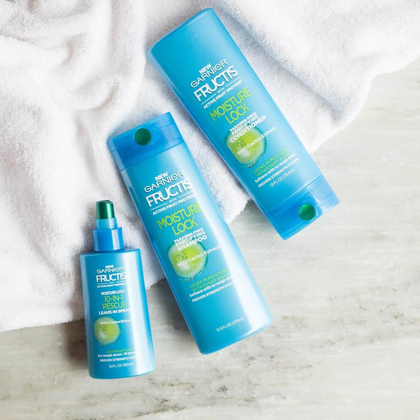 Garnier Fructis Moisture Lock Shampoo, Fructis Moisture Lock Conditioner, and 10-in-1 Rescue Leave-In Spray on grey marble and a white bath towel.